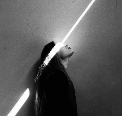 A black-and-white photograph of a woman leaning against a wall, her face lit by a single diagonal strip of light.