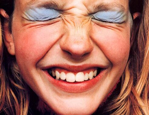 A close-up of a laughing face with both eyes screwed tightly shut and blue eyeshadow on the lids.